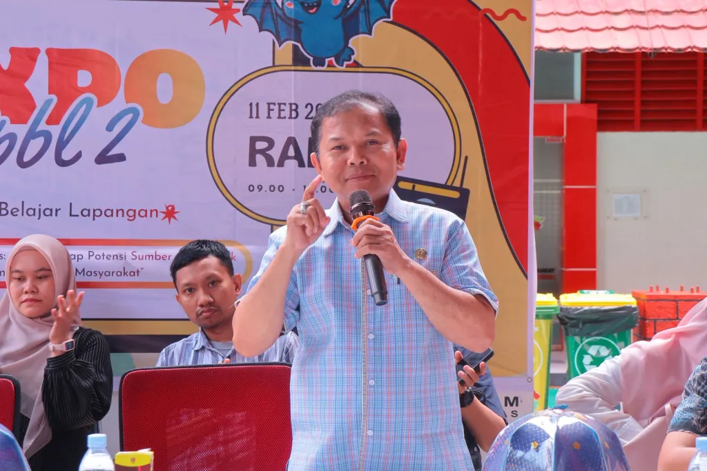 Seorang pria berbicara menggunakan mikrofon di acara kampus dengan latar belakang spanduk kegiatan.